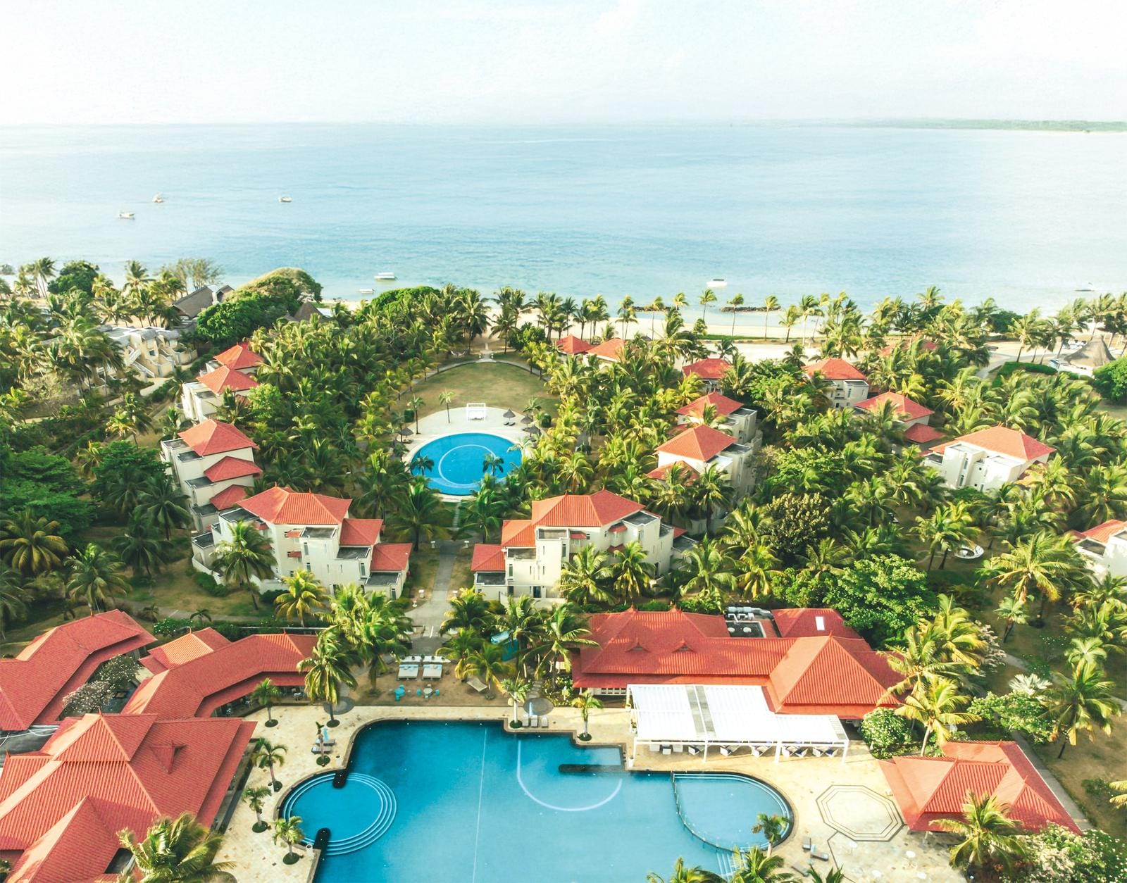 Hotelanlage am Meer mit roten Dächern, Palmen und großem Pool, entspannte Urlaubsatmosphäre.