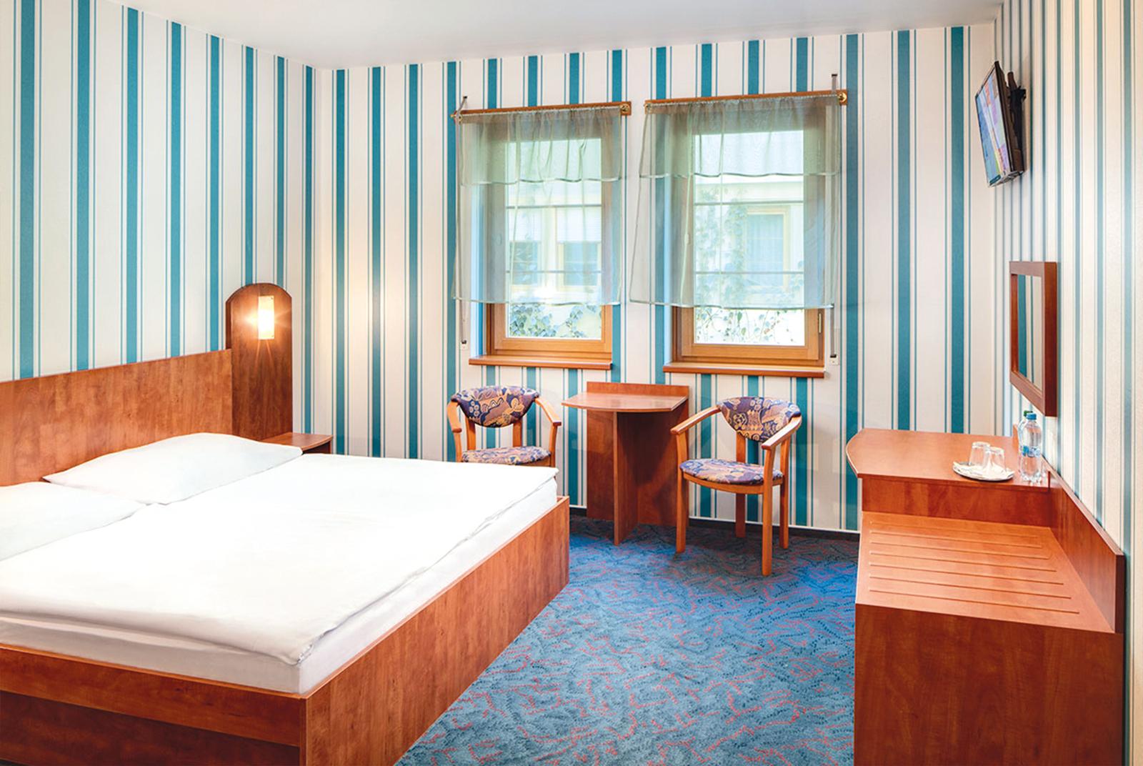 Helles Hotelzimmer mit Doppelbett, Holzoptik, blau-weiß gestreifte Wände, zwei Fenster.