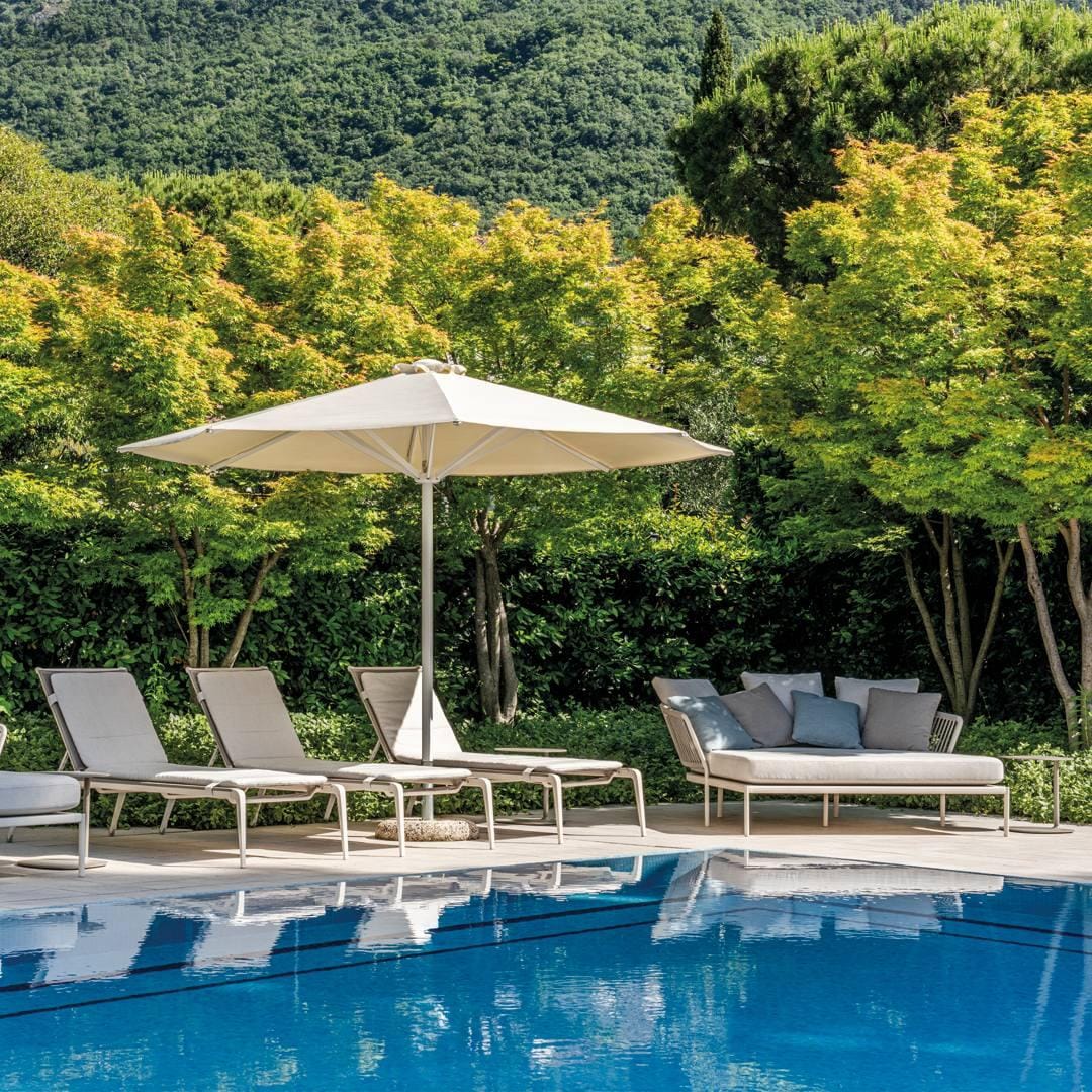 Hotelpool mit weißen Liegen, Sonnenschirm und grüner, bewaldeter Kulisse im Hintergrund.