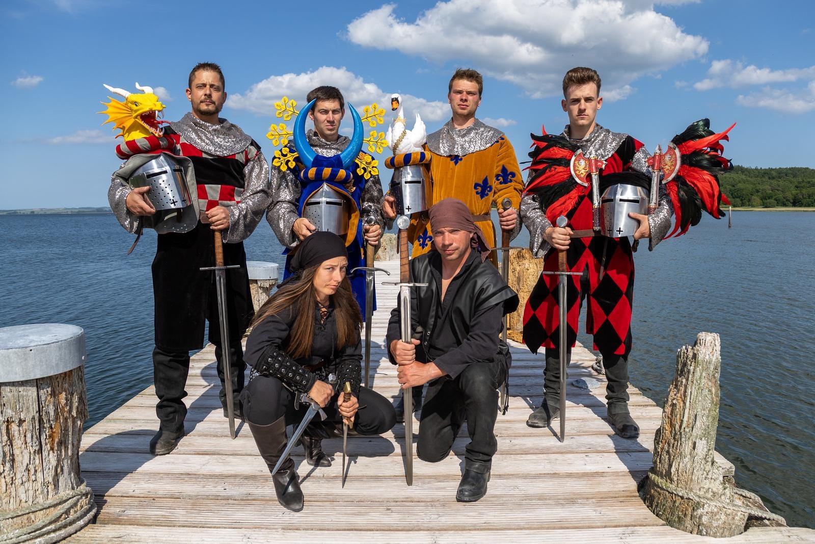Gruppe in mittelalterlichen Kostümen posiert auf Steg am See unter blauem Himmel.