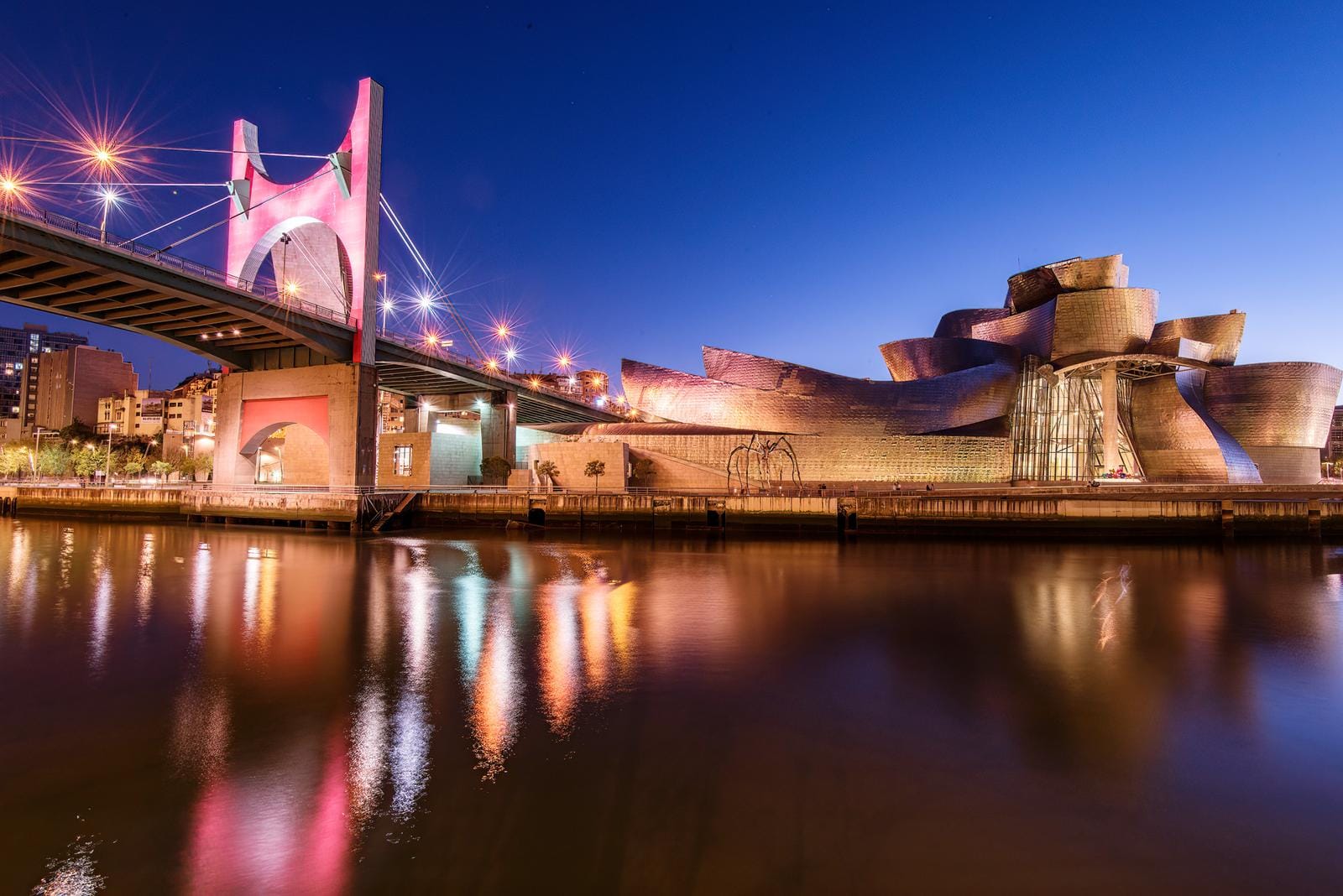 Guggenheim-Museum, Bilbao, im Abendlicht neben roter Brücke; reflektiert im Fluss.