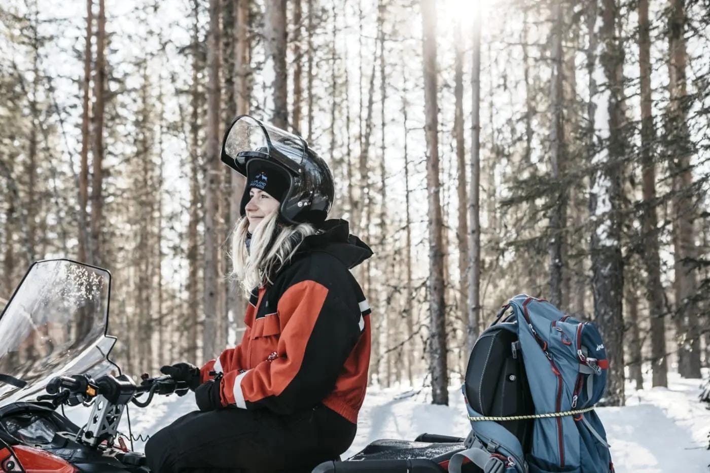 Person auf Schneemobil im winterlichen Wald mit Sonnenlicht im Hintergrund.