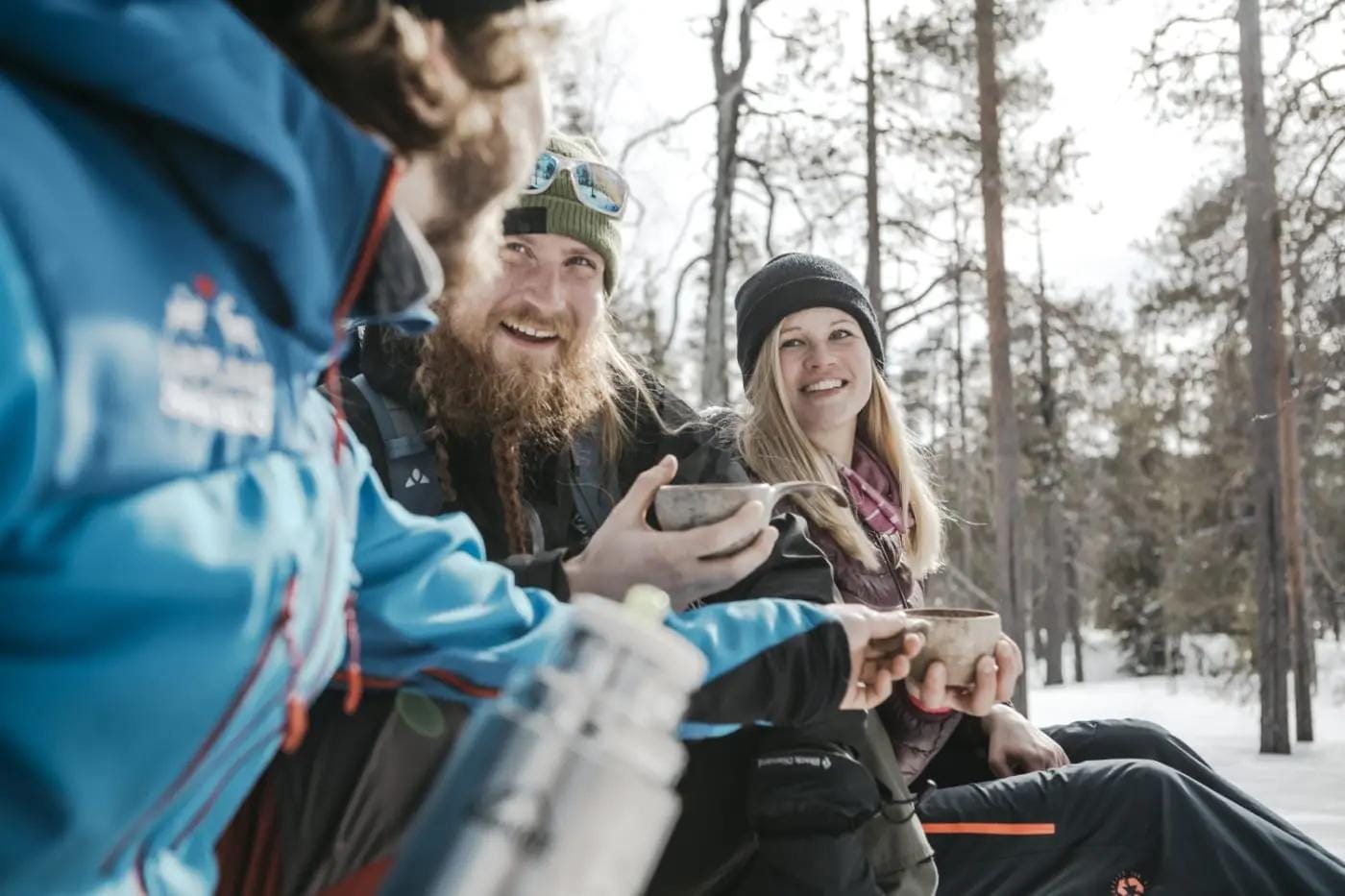 Menschen lachen im verschneiten Wald, gekleidet in Winterjacken mit Thermosflasche.