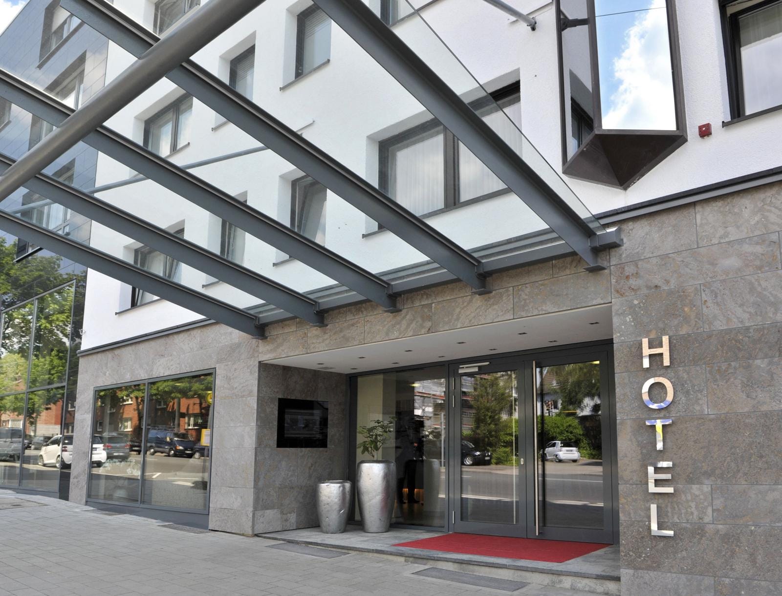 Moderne Hotelfassade mit Glasvordach und roten Willkommensmatten.