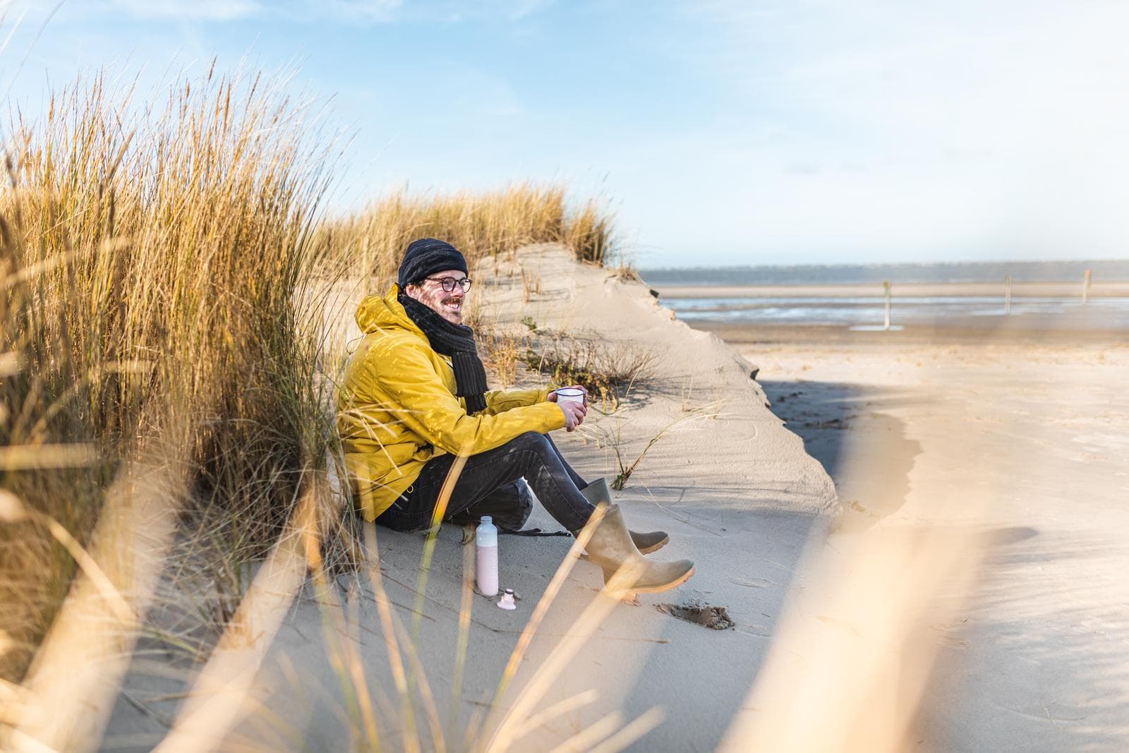 Mann in gelber Jacke sitzt lächelnd in Sanddünen am Strand, klarer blauer Himmel.