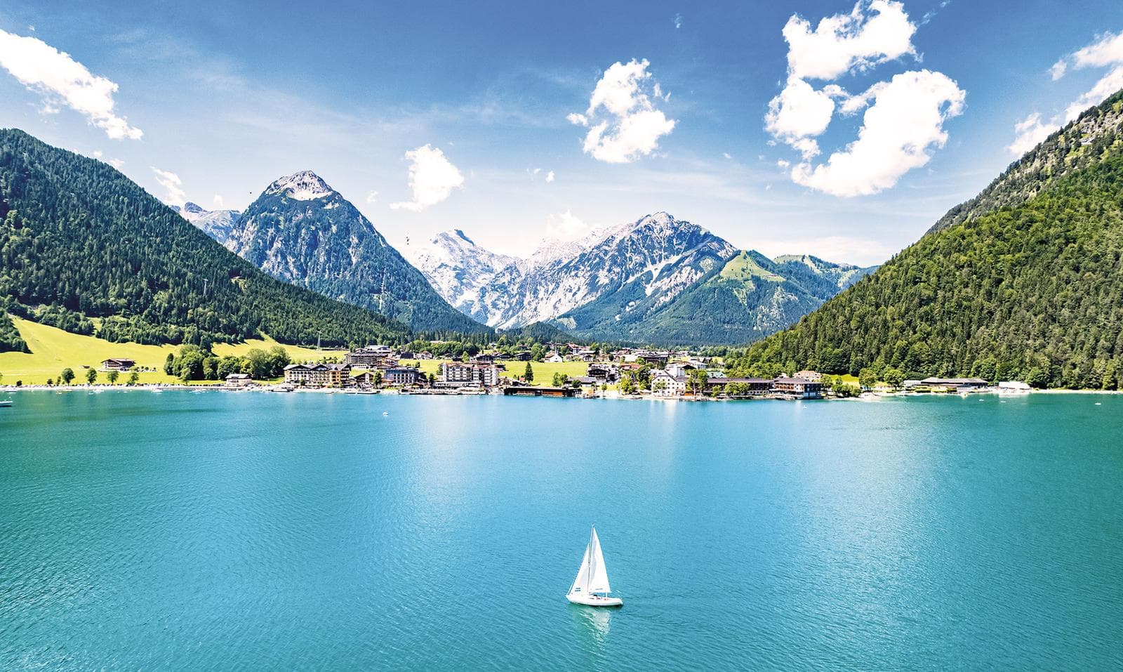 Azurblauer See mit Segelboot, umgeben von grünen Bergen und alpiner Ortschaft.