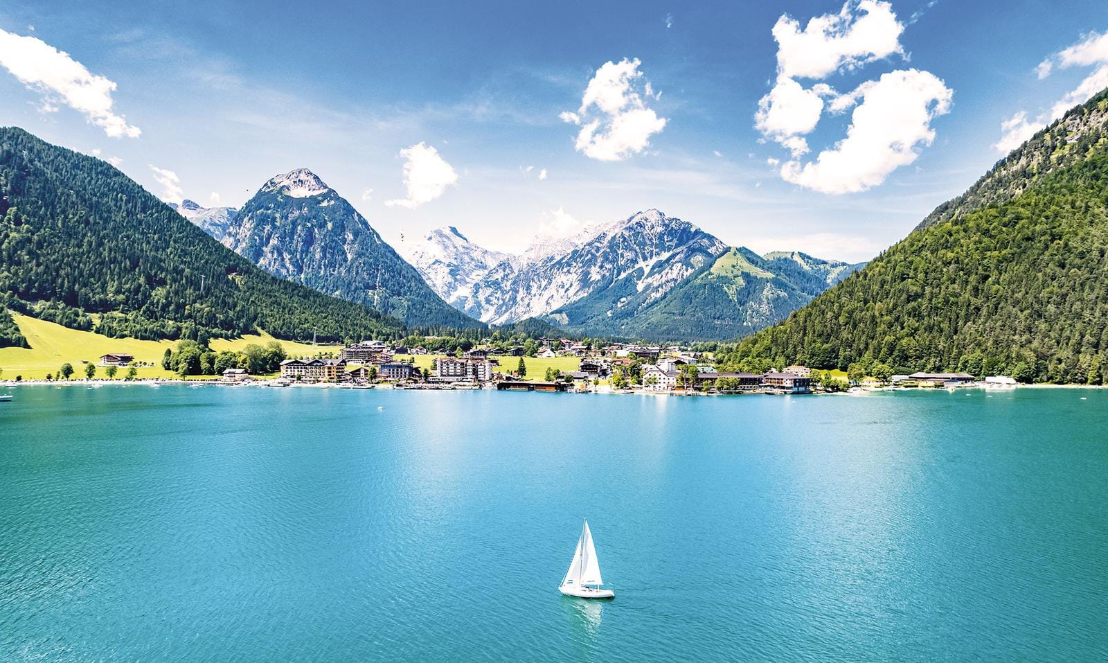 Azurblauer See mit Segelboot, umgeben von grünen Bergen und alpiner Ortschaft.