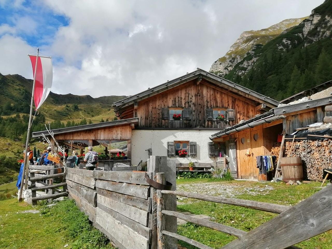 Idyllische Berghütte in den Alpen, mit rot-weißer Fahne und bewaldeten Hängen.