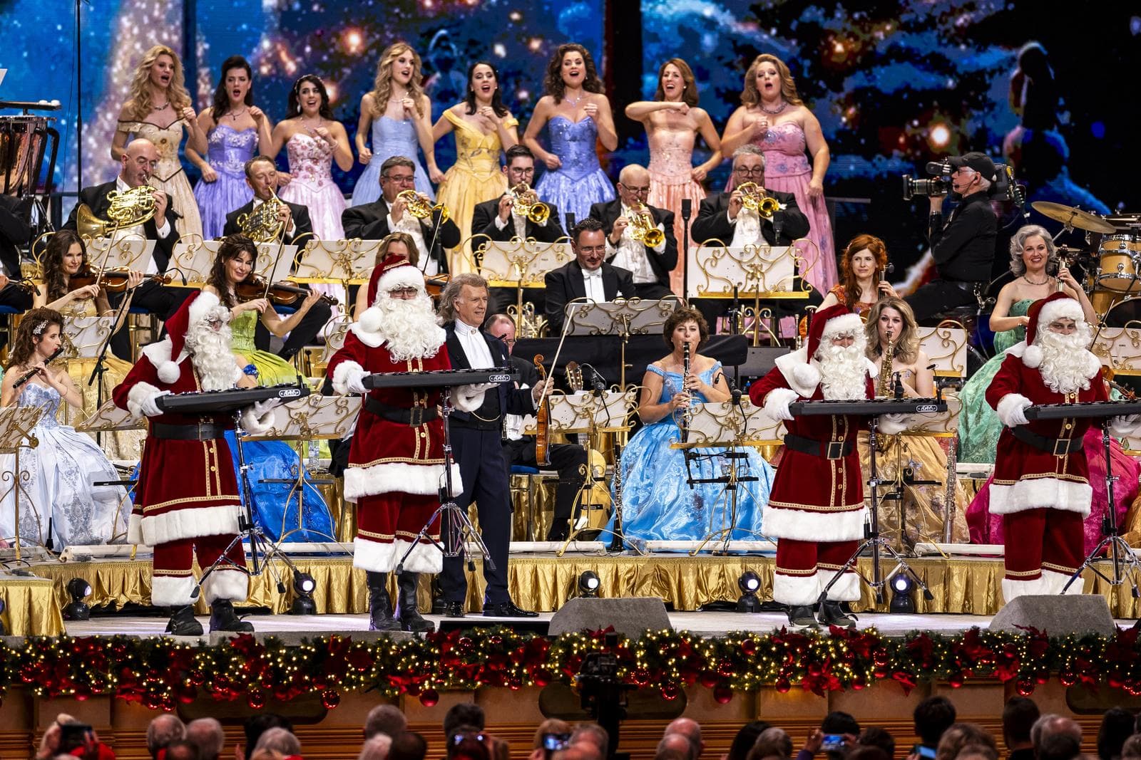 Konzertszene mit Orchester, Chorsängern in bunten Kleidern und vier Weihnachtsmännern.