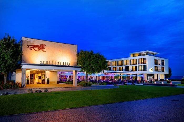 Modernes Schlosshotel mit beleuchteter Außenterrasse, umgeben von gepflegter Rasenfläche.
