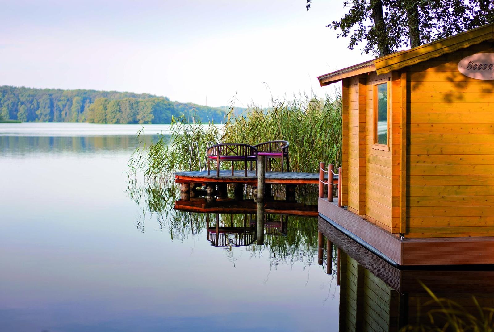 Holzhaus am See mit Anlegesteg und Sitzbank, umgeben von Schilf und ruhigem Wasser.