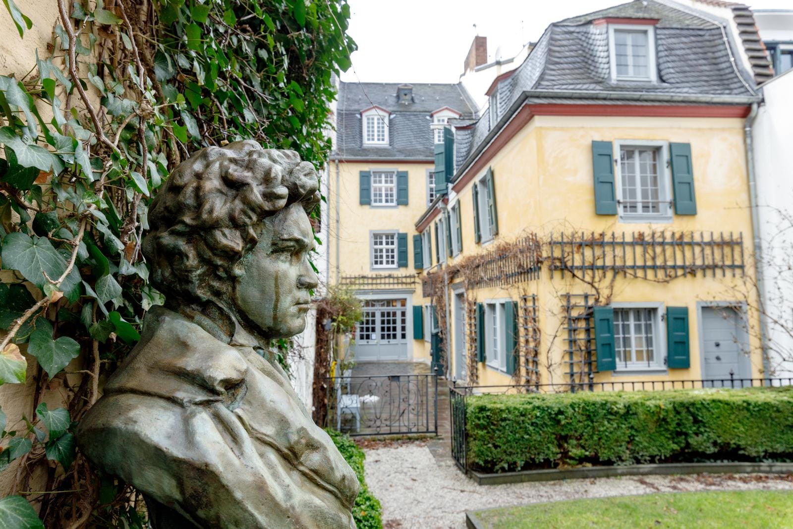 Büste vor gelbem Haus mit grünen Fensterläden, umrankt von Efeu; Bonn, Deutschland.