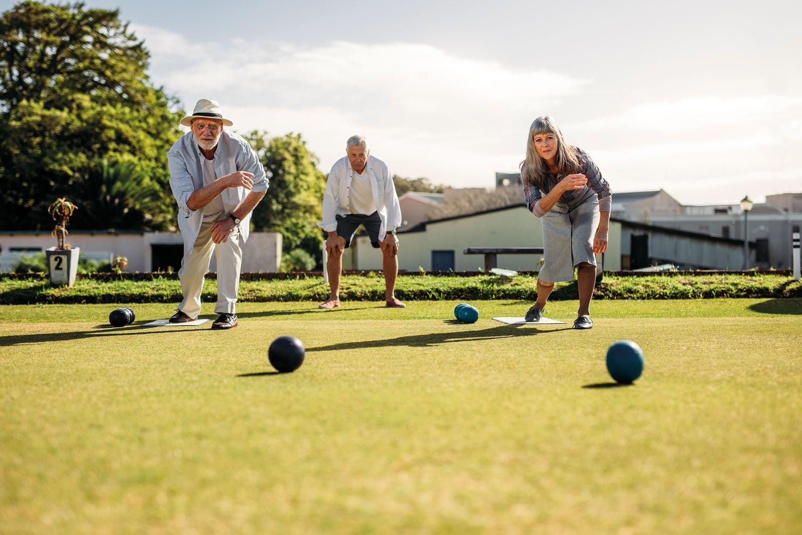 Drei Personen spielen Bowls auf einem grünen Rasenplatz bei sonnigem Wetter.