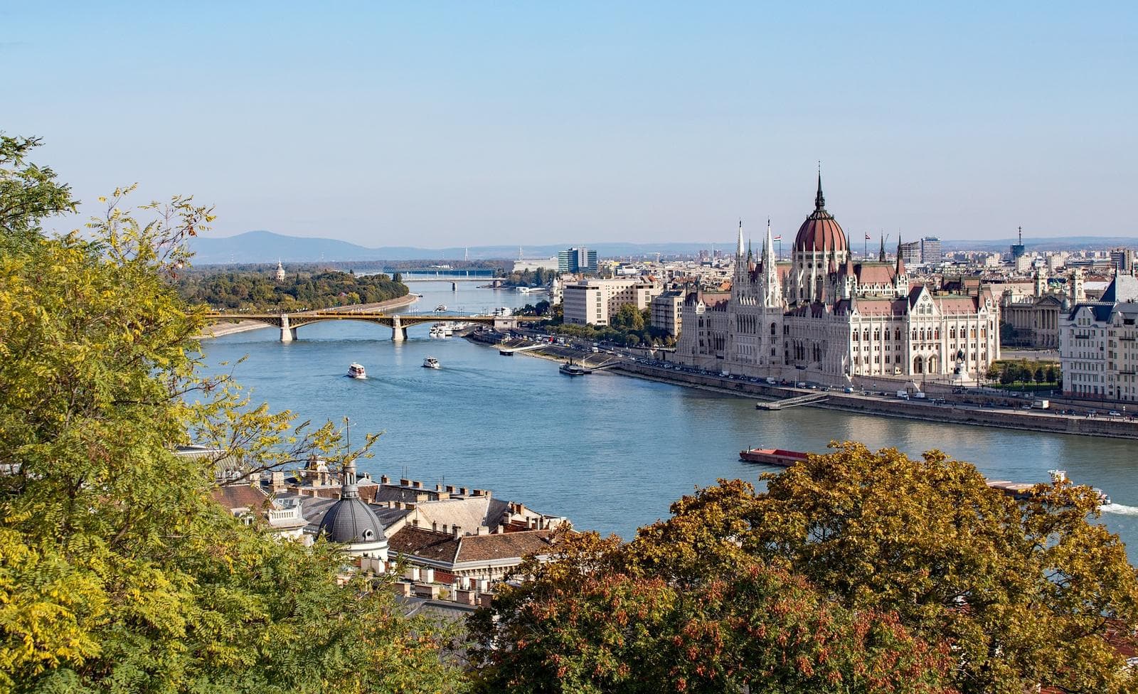 Blick auf Budapest mit der Donau im Vordergrund, Brücken und Parlament erstrahlen sonnig.