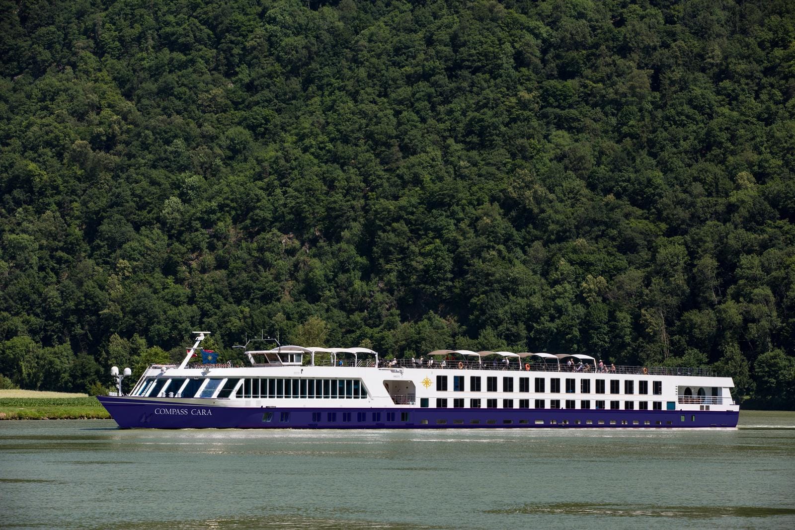 Ein weiß-lila Flusskreuzfahrtschiff fährt an einem bewaldeten Flussufer entlang.
