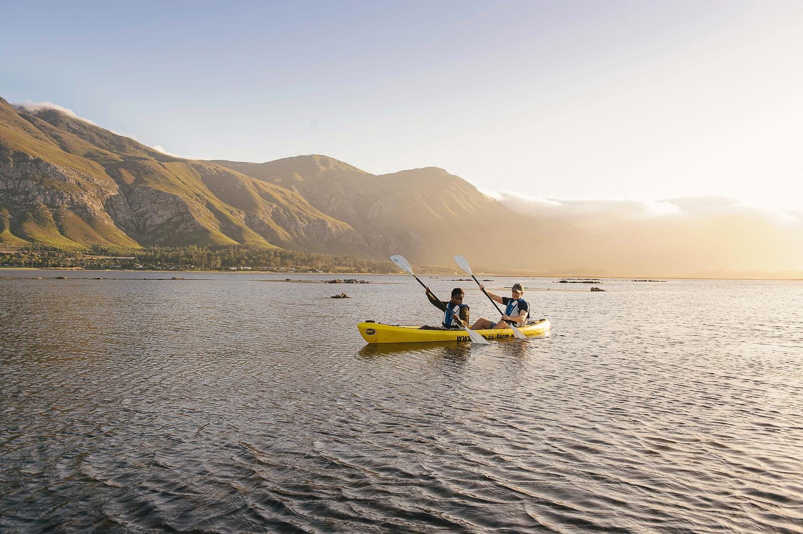 Zwei Menschen im gelben Kajak auf ruhigem See vor grĂĽnen Bergen bei Sonnenuntergang.