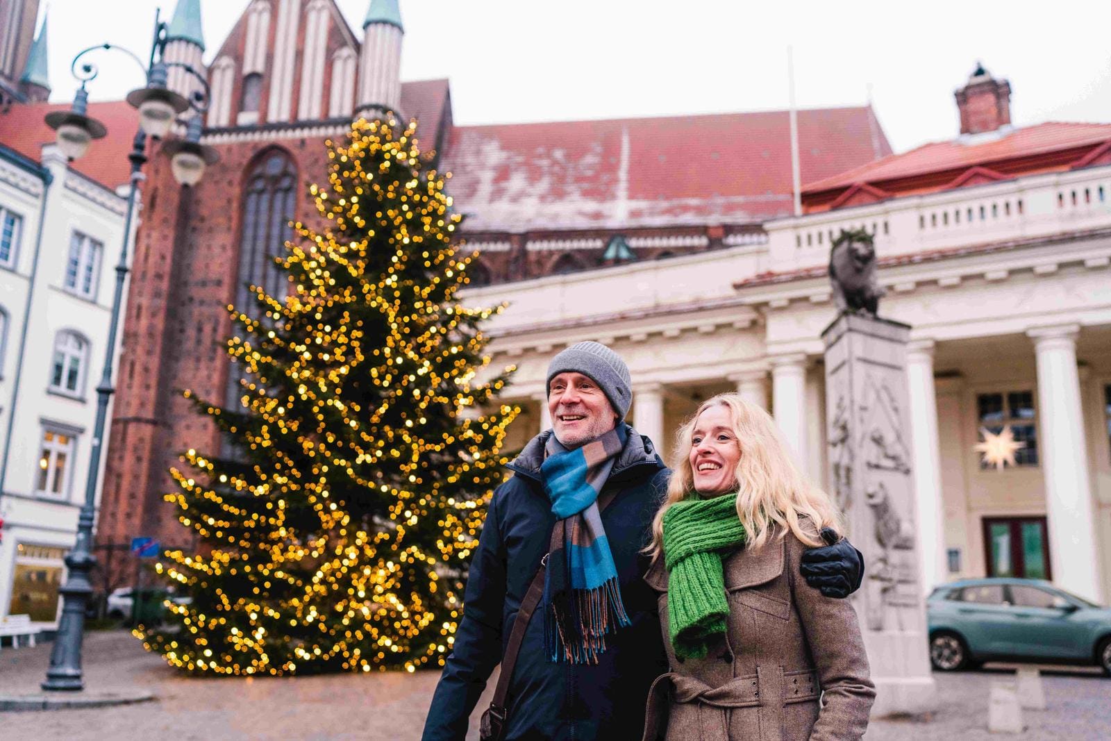 Paar vor beleuchtetem Weihnachtsbaum, historische Gebäude im Hintergrund, winterliche Stimmung.