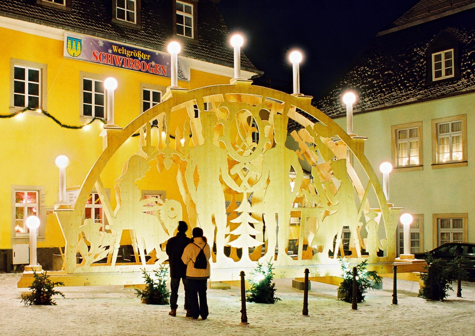 Leuchtender Holz-Schwibbogen vor gelbem Gebäude in winterlicher, abendlicher Szene.