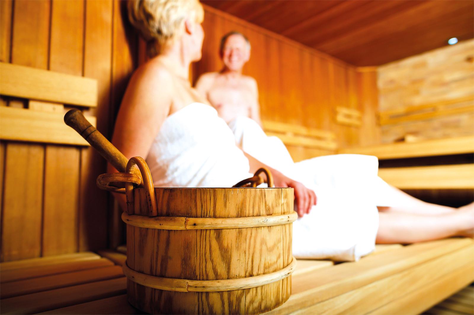 Zwei entspannte Personen in einer warmen Holzsauna mit Saunaeimer im Vordergrund.