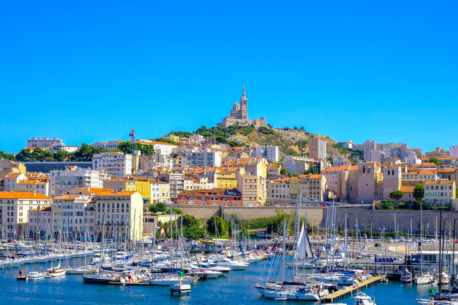 Hafen von Marseille mit Blick auf Notre-Dame de la Garde unter strahlend blauem Himmel.