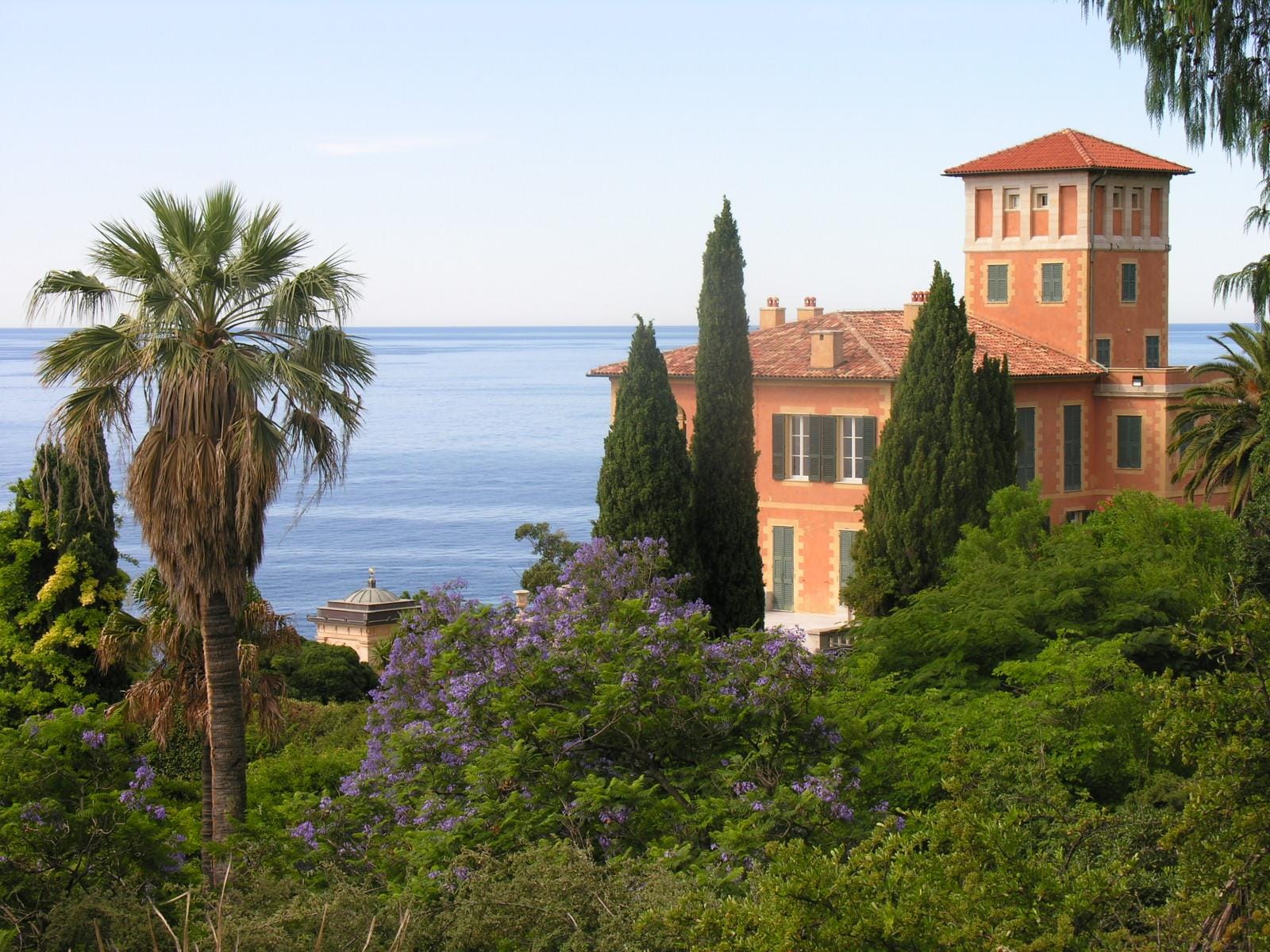 Mediterrane Villa mit roten Dächern, Palmen und Meerblick vor blühender Flora.