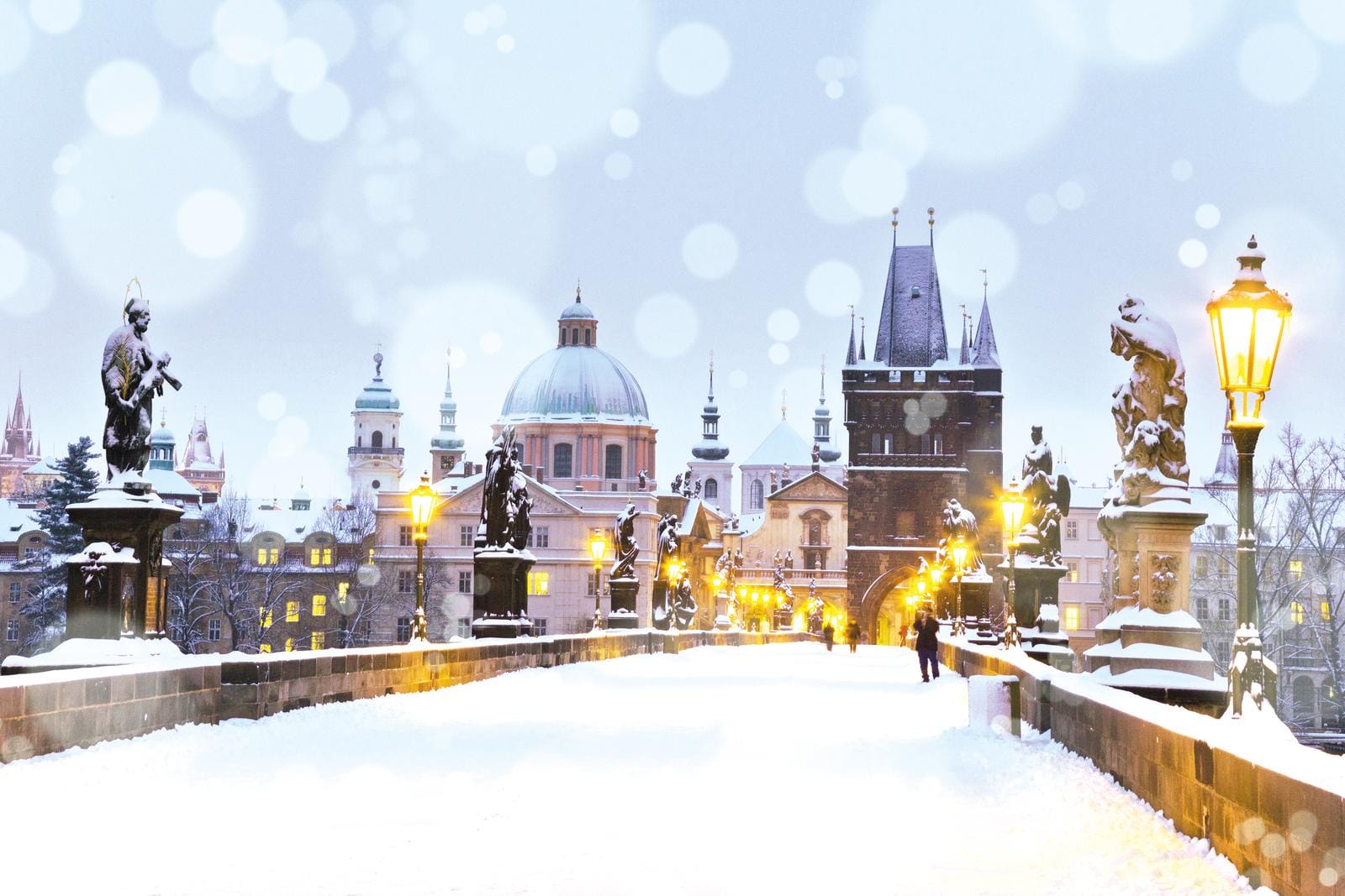 Schneebedeckte Karlsbrücke in Prag mit leuchtenden Laternen und Architektur im Hintergrund.