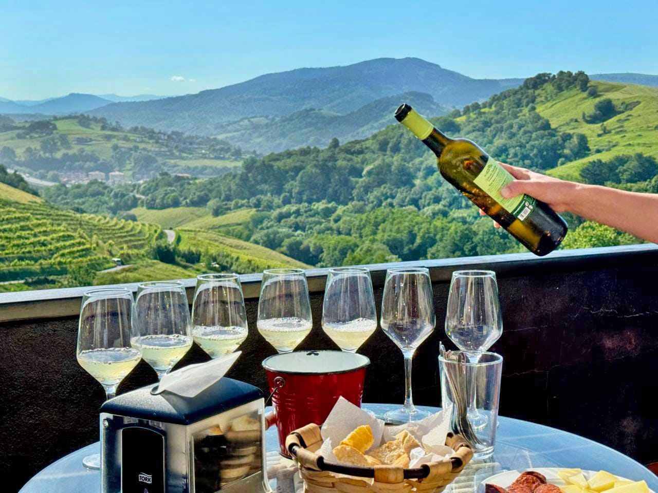 Tisch mit Wein und Snacks, Aussicht auf grüne Hügel und Weinberge bei strahlendem Sonnenschein.