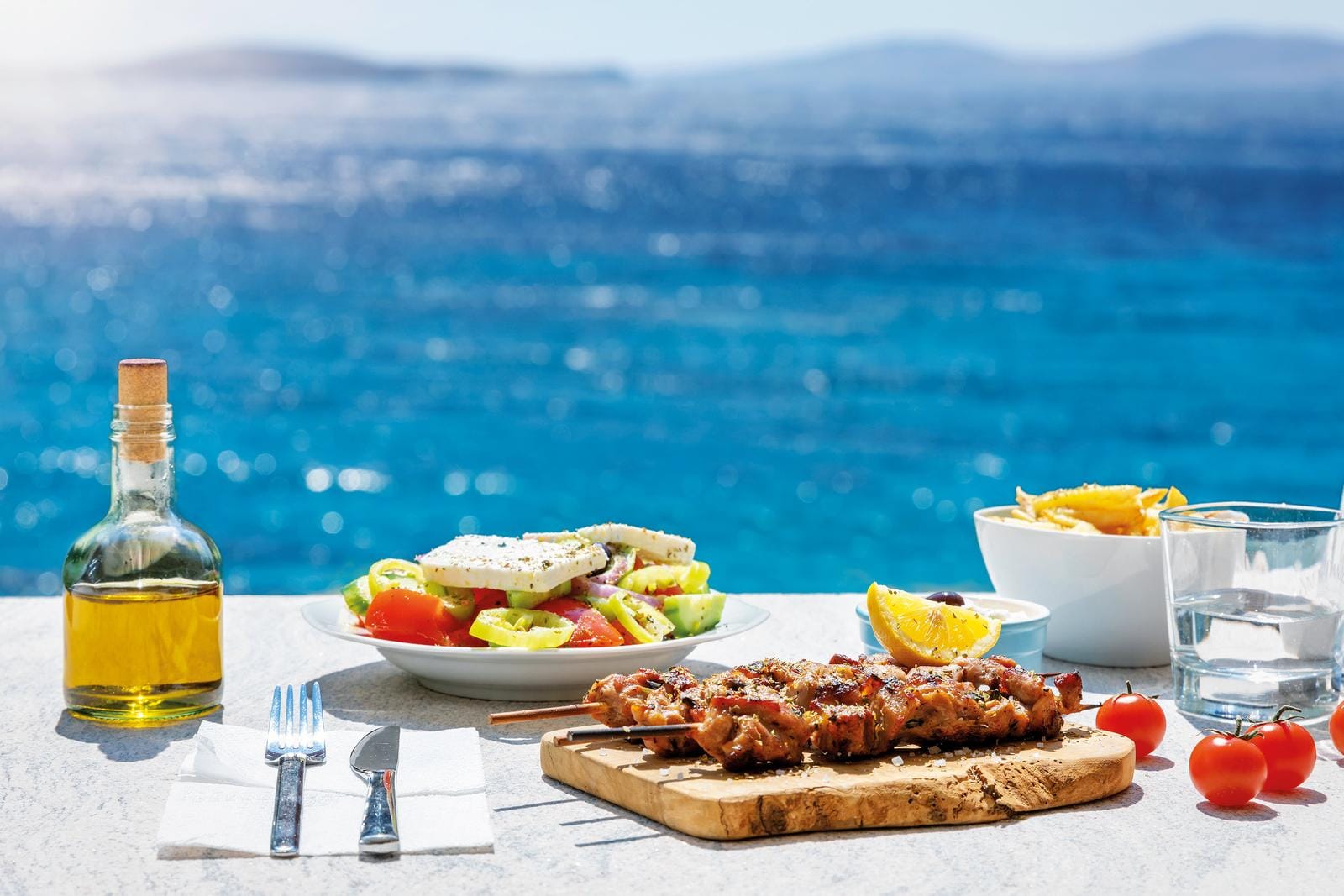 Griechisches Mittagessen mit Salat und Fleischspieß am Meer, Sonnenschein im Hintergrund.