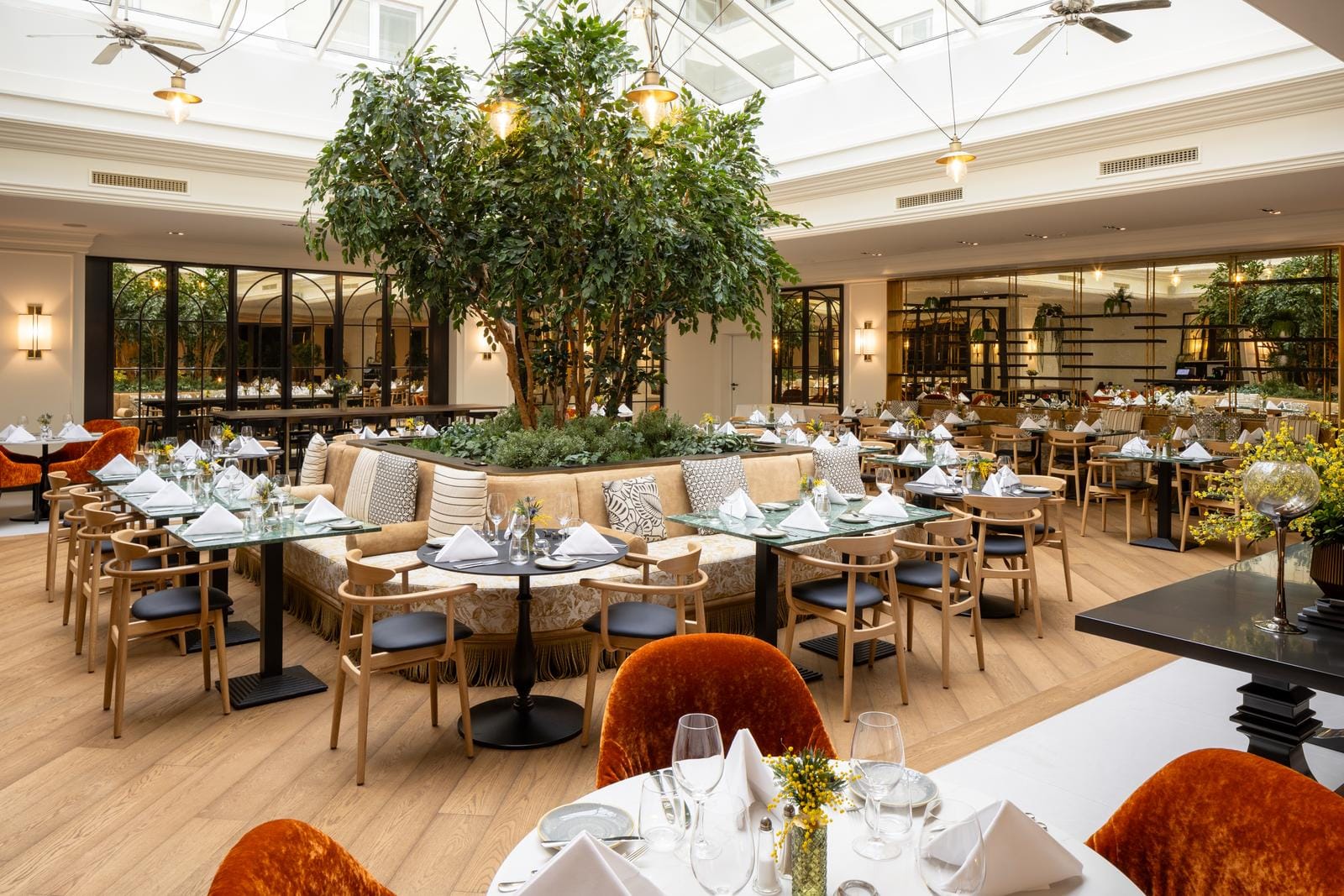 Elegantes Hotelrestaurant mit viel Grün, Holzböden, Glasdach und stilvollen Möbeln.