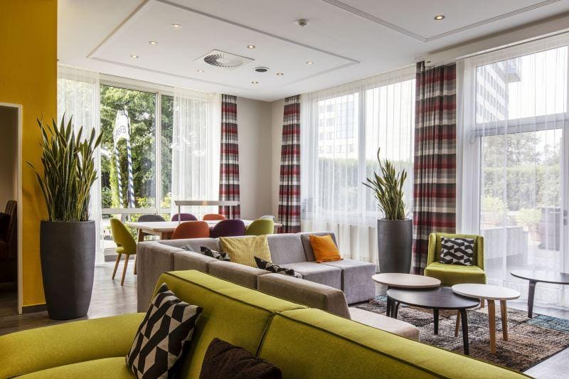 Moderne Hotel-Lounge mit bunten Sofas, großen Fenstern und Pflanzen.