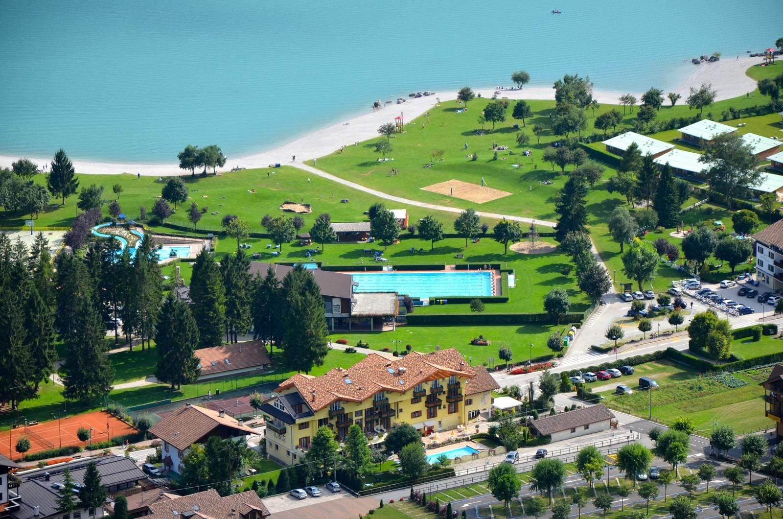 Hotelanlage am See mit Pool, Tennisplatz und grüner Liegewiese umgeben von Bäumen.