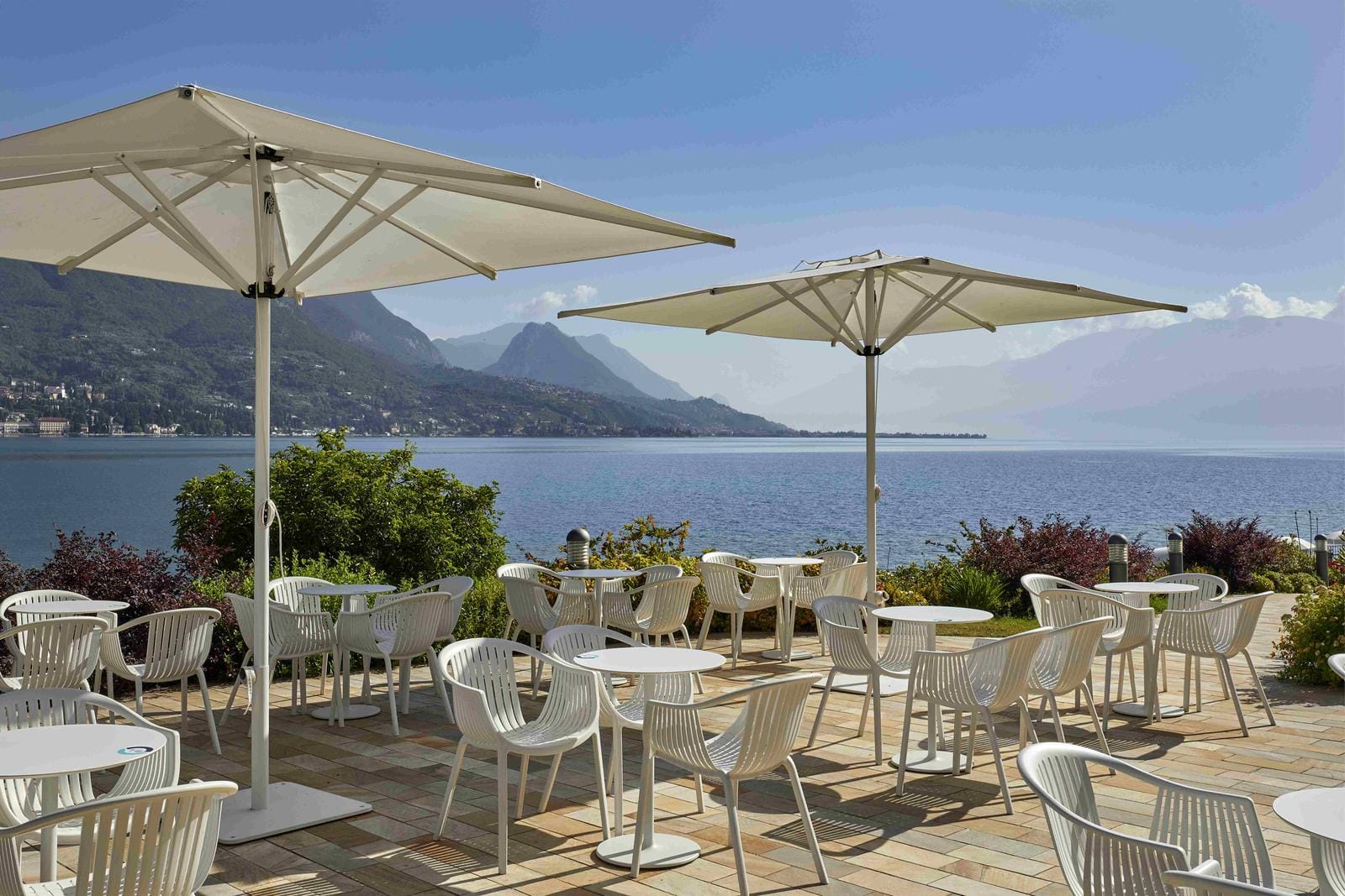 Terrasse mit weißen Tischen und Stühlen, Blick auf einen See und umliegende Berge.