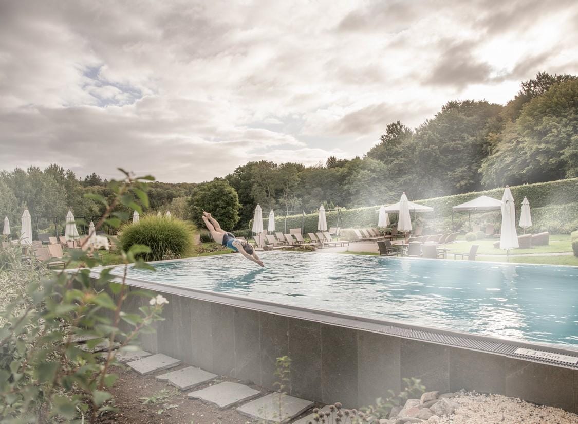 Mann springt in modernem Hotelpool mit Liegen, Sonnenschirmen, umgeben von Bäumen.