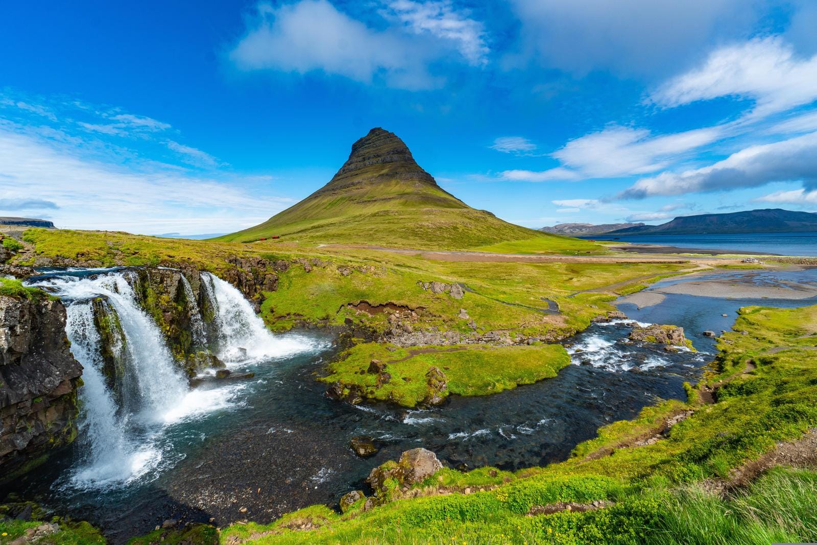 Kirkjufell, Island: GrĂĽner HĂĽgel mit Wasserfall und blauem Himmel im Hintergrund.