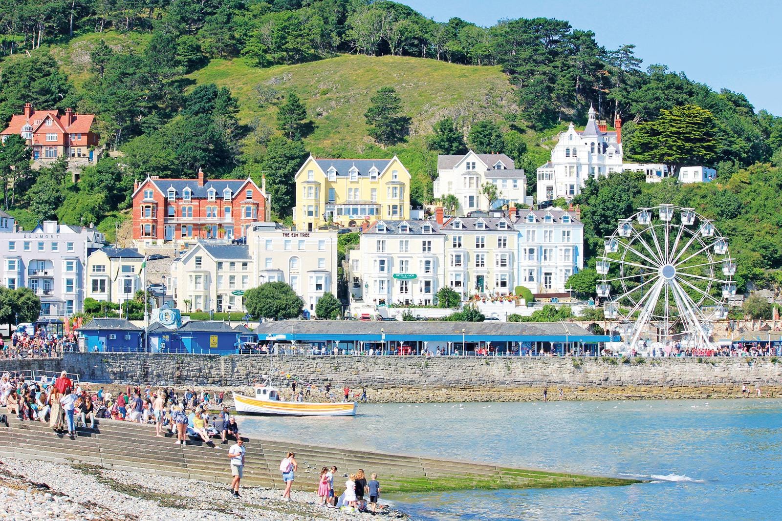 Bunte Häuserreihe am Hang, Riesenrad und Küste, lebhaftes Sommerflair in Llandudno.