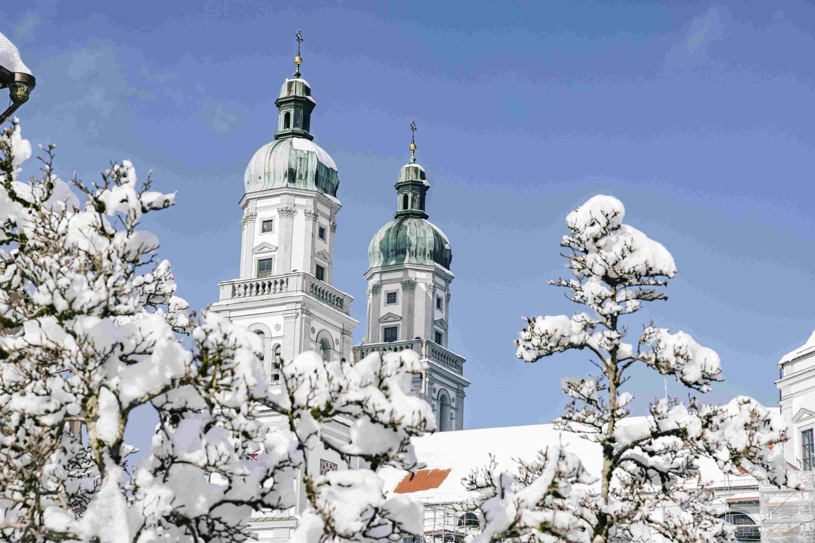 Verschneite Doppeltürme einer barocken Kathedrale vor blauem Himmel, verschneite Bäume.