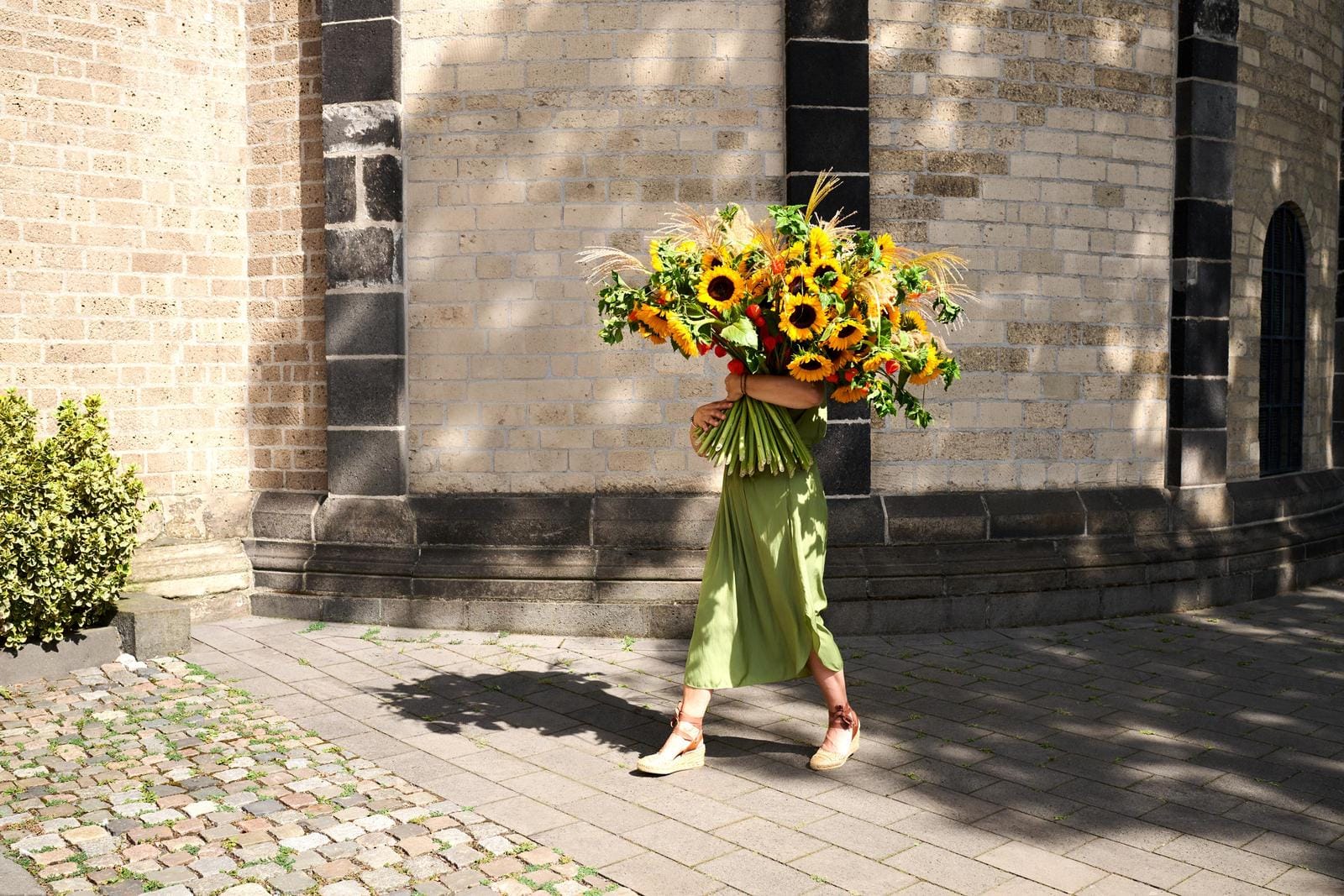 Person in grünem Kleid trägt großen Sonnenblumenstrauß vor Backsteinwand.