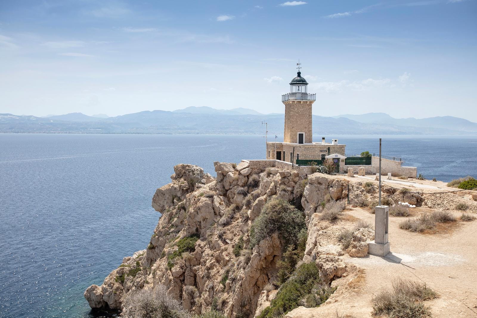 Leuchtturm auf felsiger Küste mit Blick auf das blaue Meer und entfernte Berge in Griechenland.