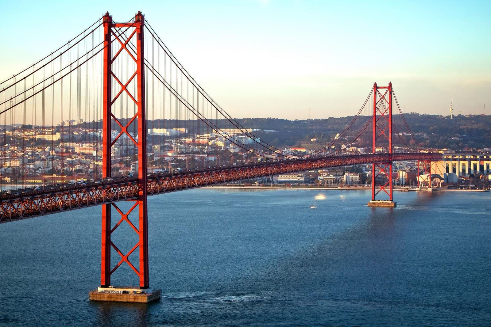 Rote Hängebrücke über blauem Fluss bei Lissabon, Portugal, vor urbaner Landschaft.