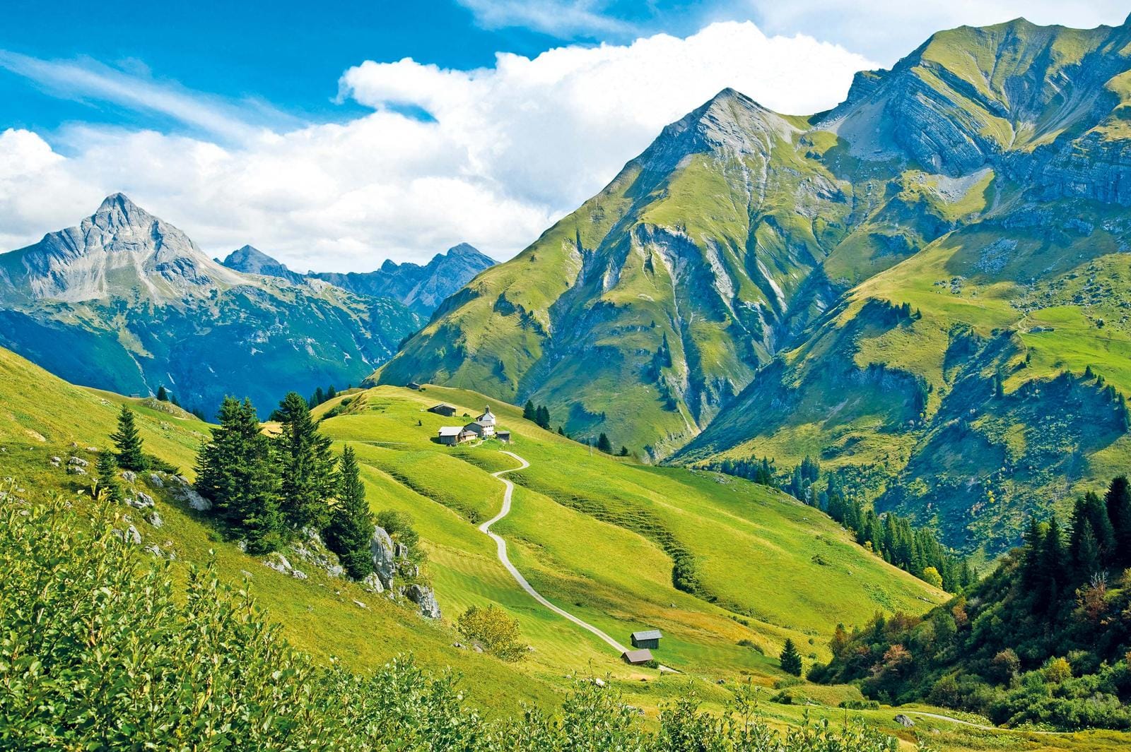 Grüne Almlandschaft mit Hütten, eindrucksvolle Berge unter blauem Himmel, Österreich.