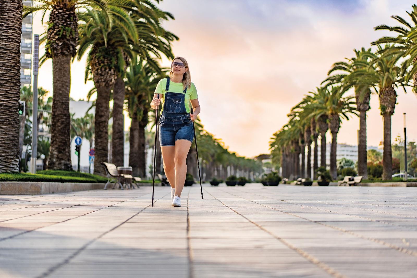 Frau beim Nordic Walking auf einer von Palmen gesäumten Promenade bei Sonnenuntergang.