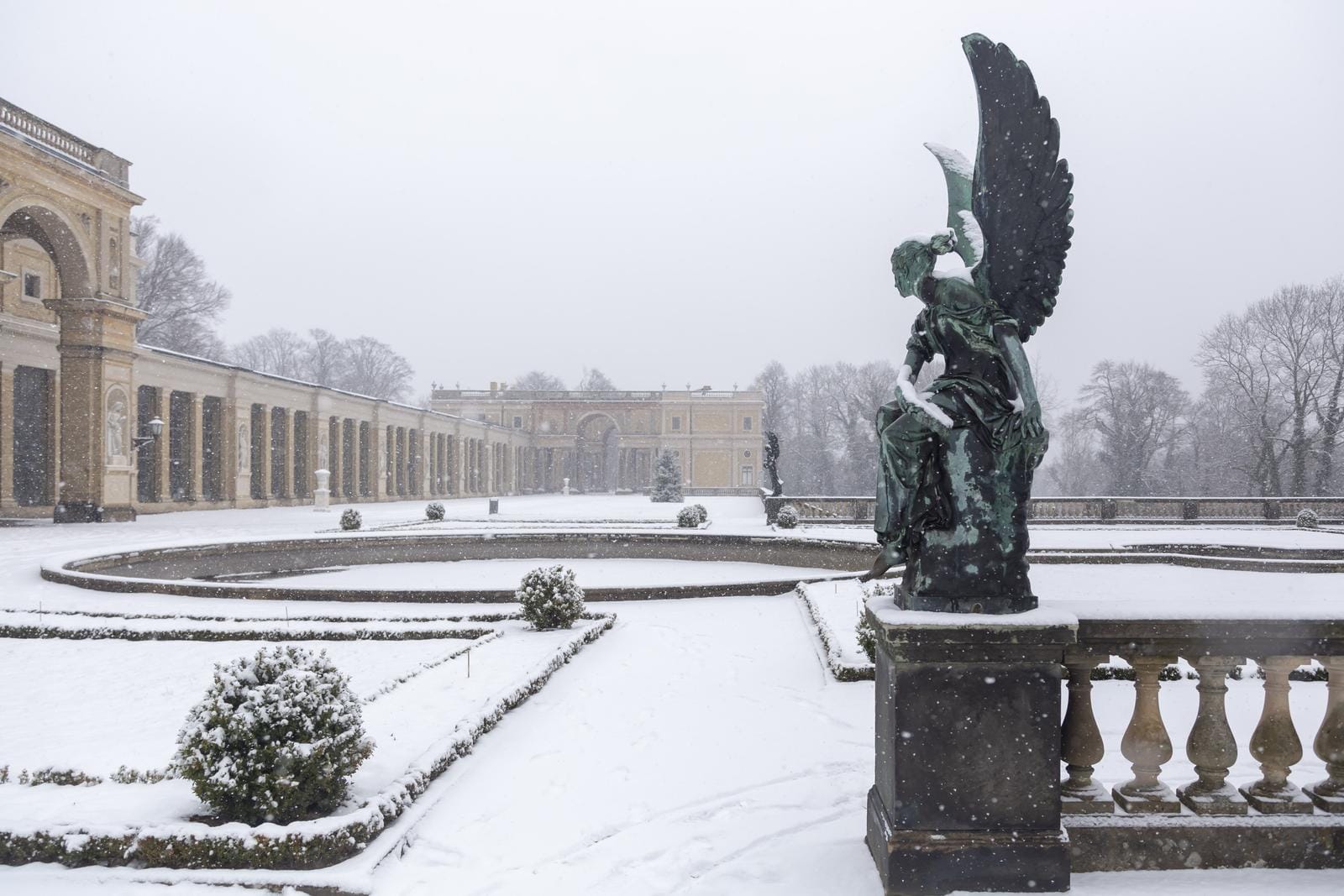 Schneebedeckter Wintergarten mit Engelstatue und historischem Gebäude im Hintergrund.