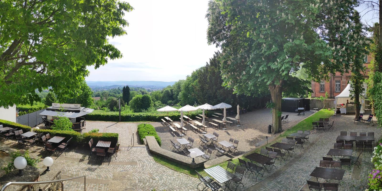Gartenrestaurant mit Terrassenmöbeln, Sonnenschirmen und weitem Blick auf grüne Hügel.