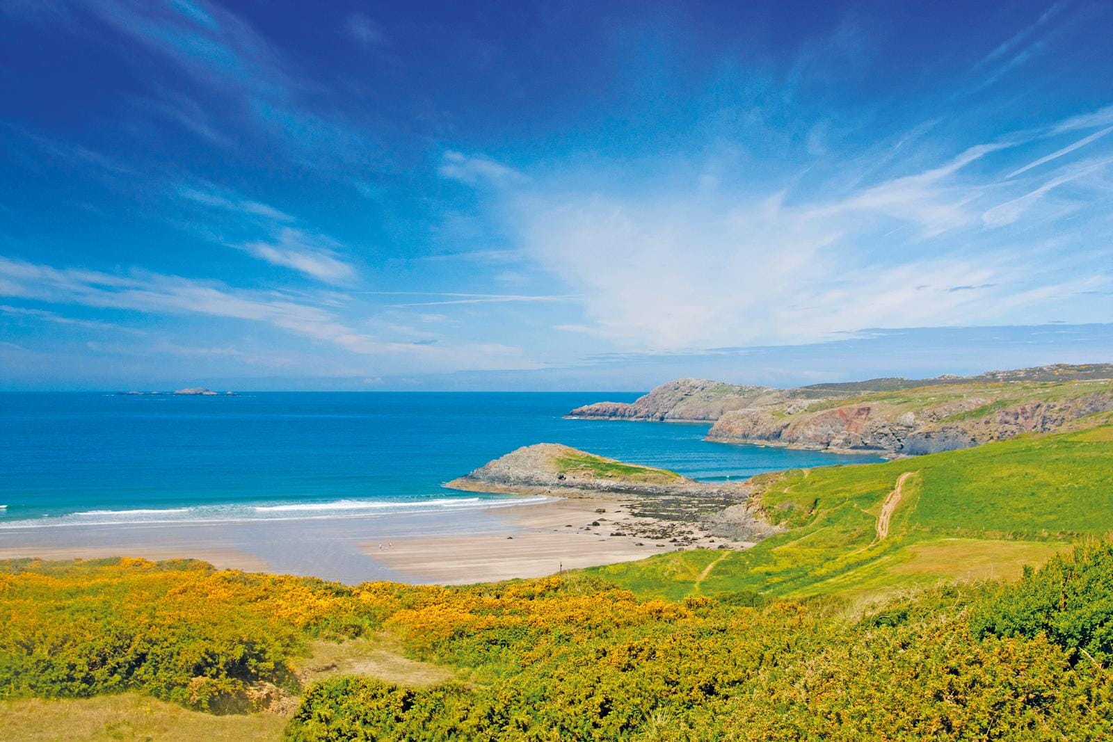 Steile Küste in Wales mit blauem Meer, Sandstrand und grünen Hügeln.