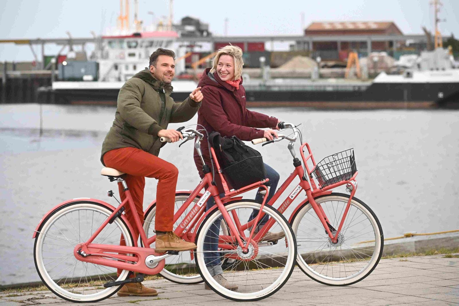 Zwei Personen radeln auf einem roten Fahrrad entlang eines Hafens.