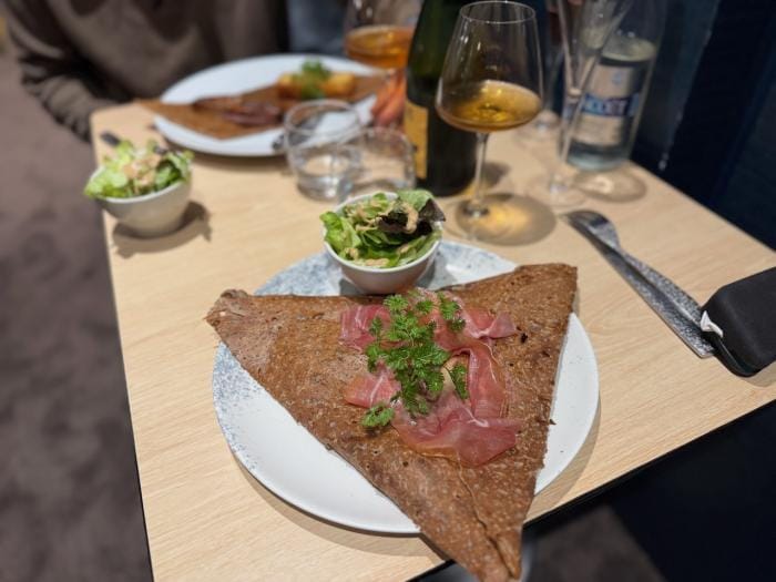 Galette mit Schinken und Kräutern, dazu Salat und Weißwein auf einem Holztisch.