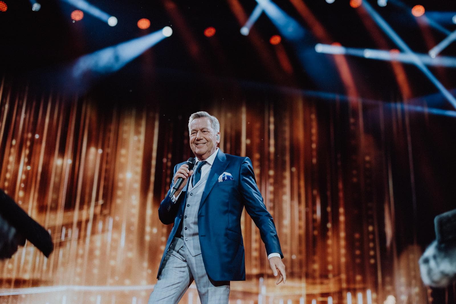 Eine Person in blauem Anzug auf Bühne mit bunten Lichtern und Mikrofon.