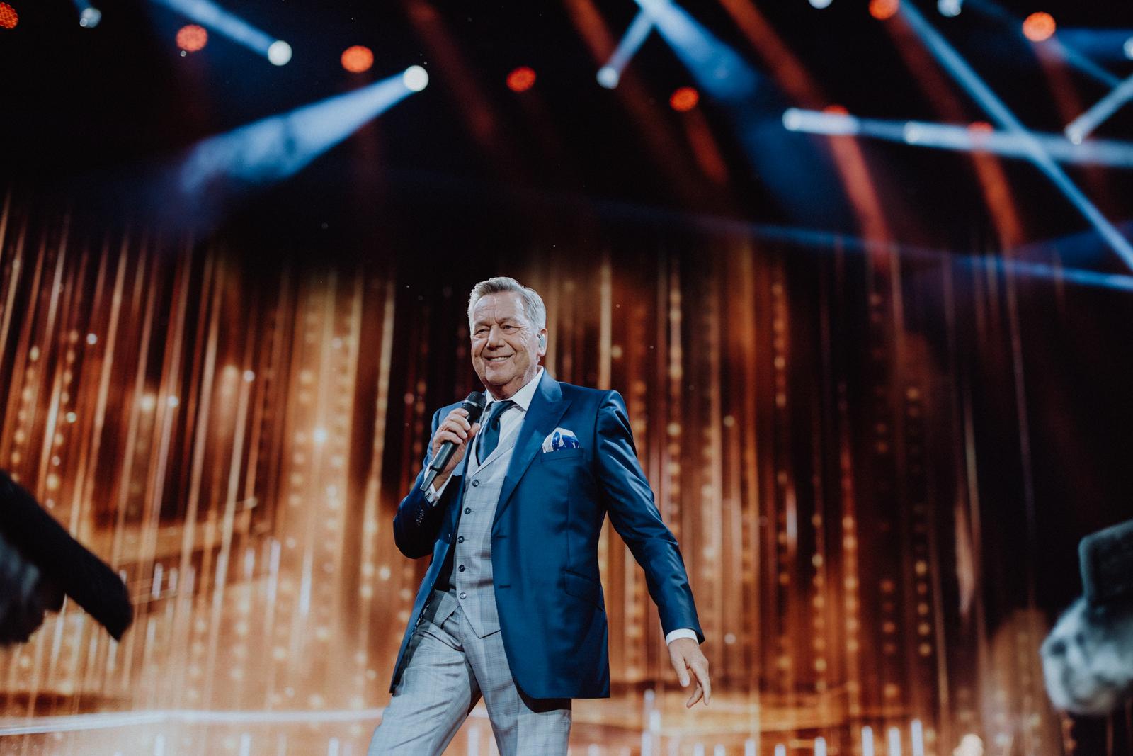 Eine Person in blauem Anzug auf Bühne mit bunten Lichtern und Mikrofon.