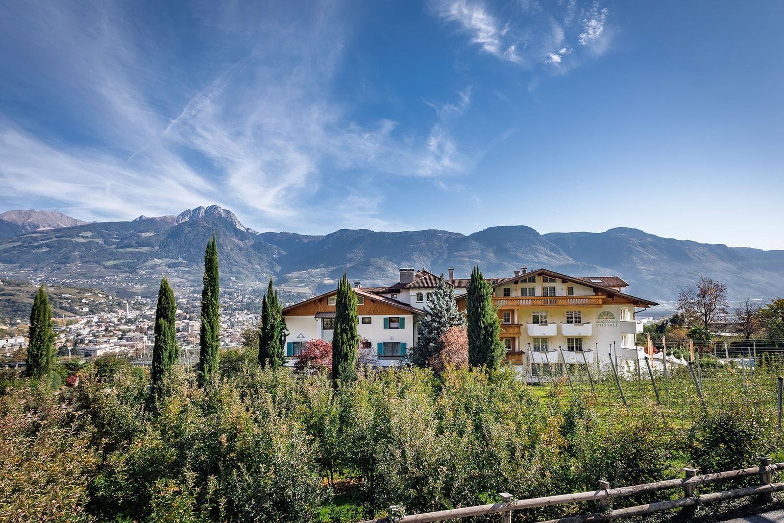 Hotel in Südtiroler Landschaft mit Zypressen, Bergen und blauem Himmel im Hintergrund.