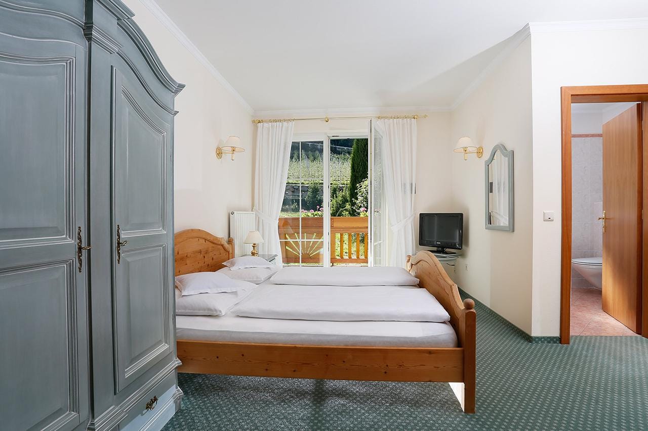 Gemütliches Hotelzimmer mit Holzmöbeln, Balkon, TV, weißem Vorhang, helles Ambiente.